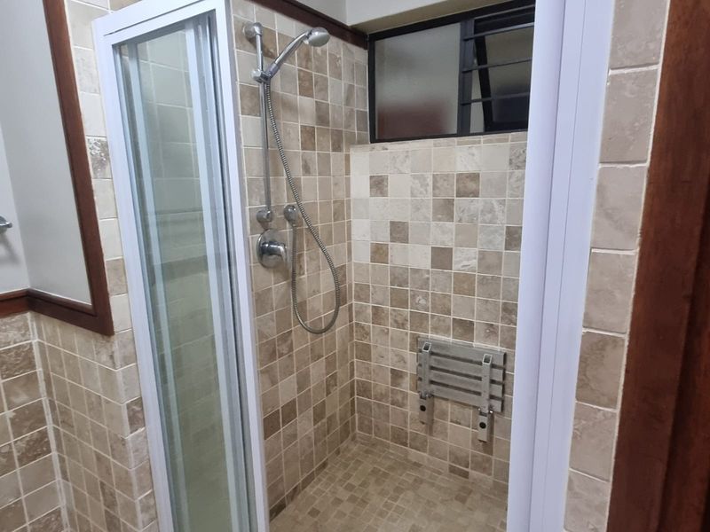 A functional bathroom shower area featuring neutral-toned wall and floor tiles, a built-in shower seat, and a louvered window for ventilation.