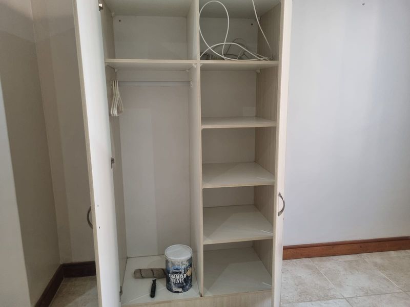 A built-in wardrobe with shelving and hanging space, featuring a light wood-grain finish and tiled flooring.