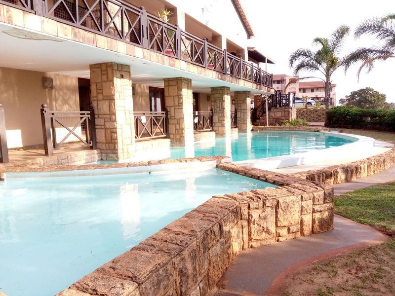 A communal swimming pool area featuring stone-clad pillars and a raised balcony with timber railings, set within a landscaped apartment complex.