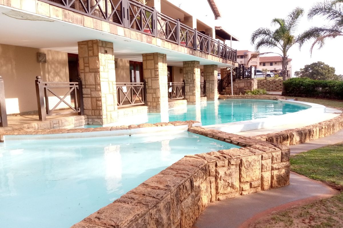 A communal swimming pool area featuring stone-clad pillars and a raised balcony with timber railings, set within a landscaped apartment complex.