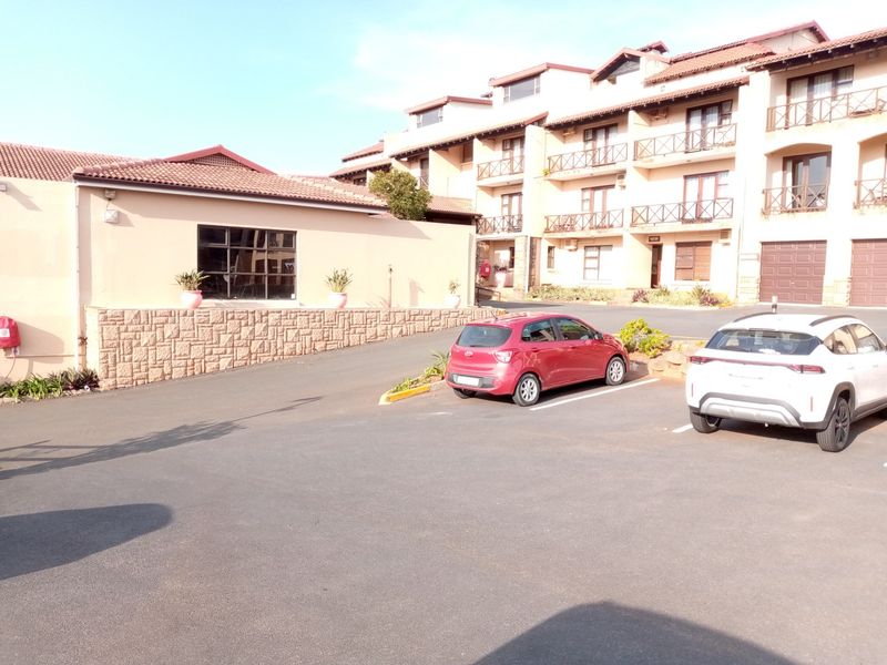 Exterior view of an apartment complex featuring a paved parking area, stone retaining wall, and multi-level residential units with balconies.