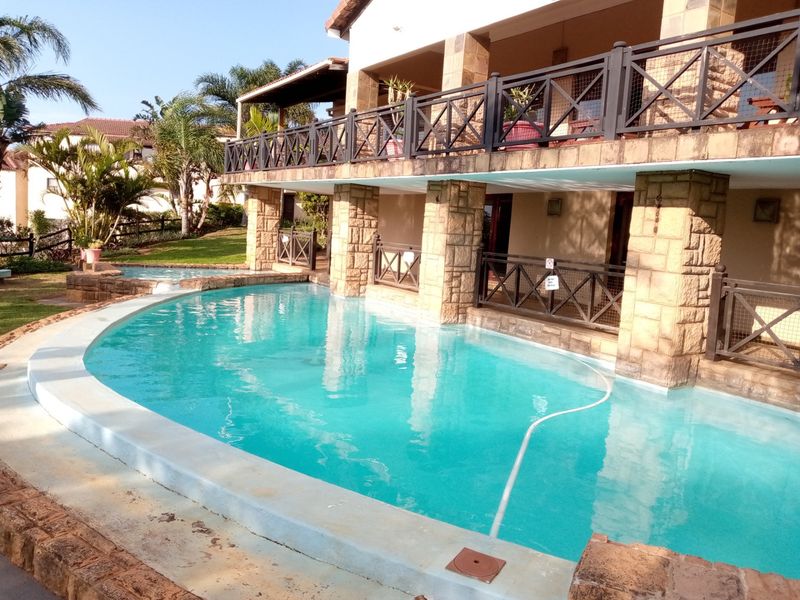 A swimming pool area featuring stone-clad pillars and a balcony with railings, set against a landscaped garden background.