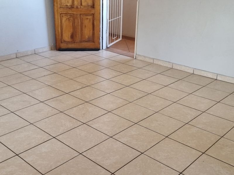 An empty living area featuring neutral ceramic floor tiles, tiled skirting, and a wooden door leading to an adjacent space.