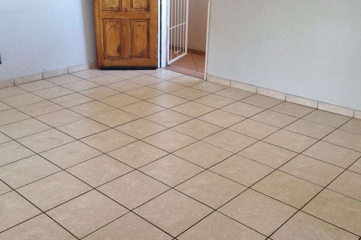 An empty living area featuring neutral ceramic floor tiles, tiled skirting, and a wooden door leading to an adjacent space.