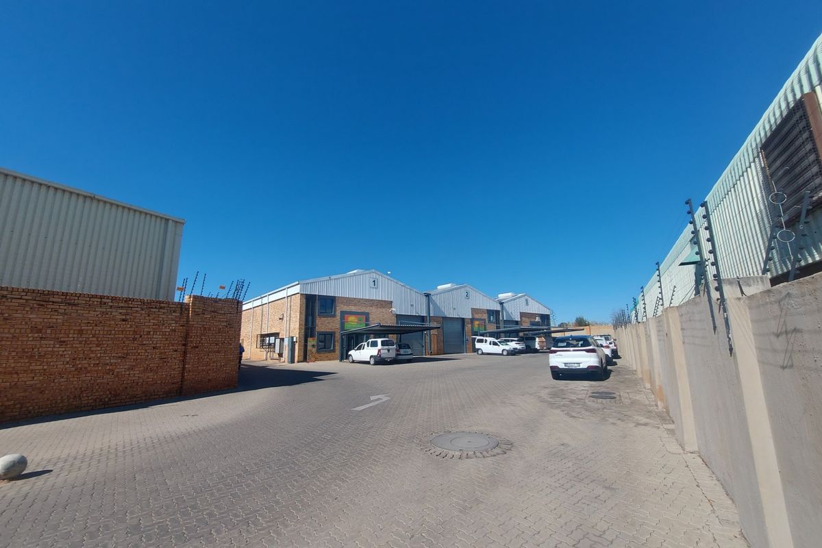 Exterior view of an industrial complex featuring multiple warehouse units with paved driveway access, perimeter brick walls, and security fencing.
