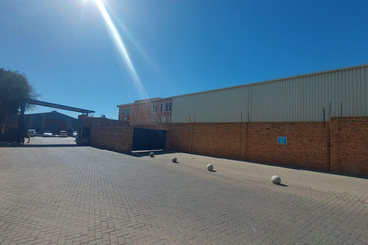 Exterior view of the industrial property featuring a paved driveway, brick perimeter wall with security fencing, and access gate.