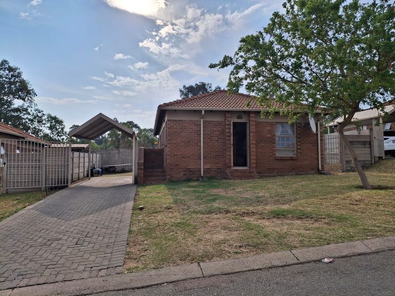 Front view of a single-story brick house featuring a tiled roof, a paved driveway with a carport, and a fenced yard.