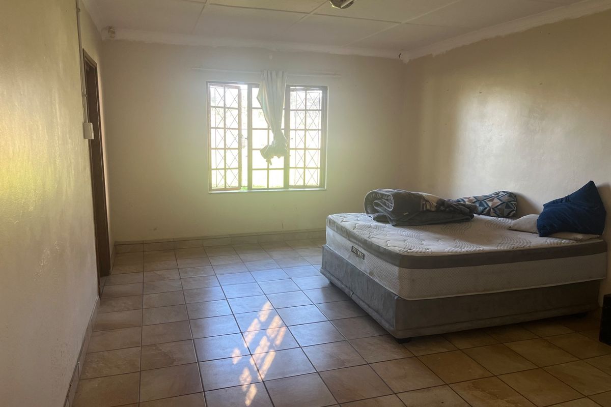 A spacious bedroom featuring tiled flooring, a large window with security bars, and a simple, functional layout.