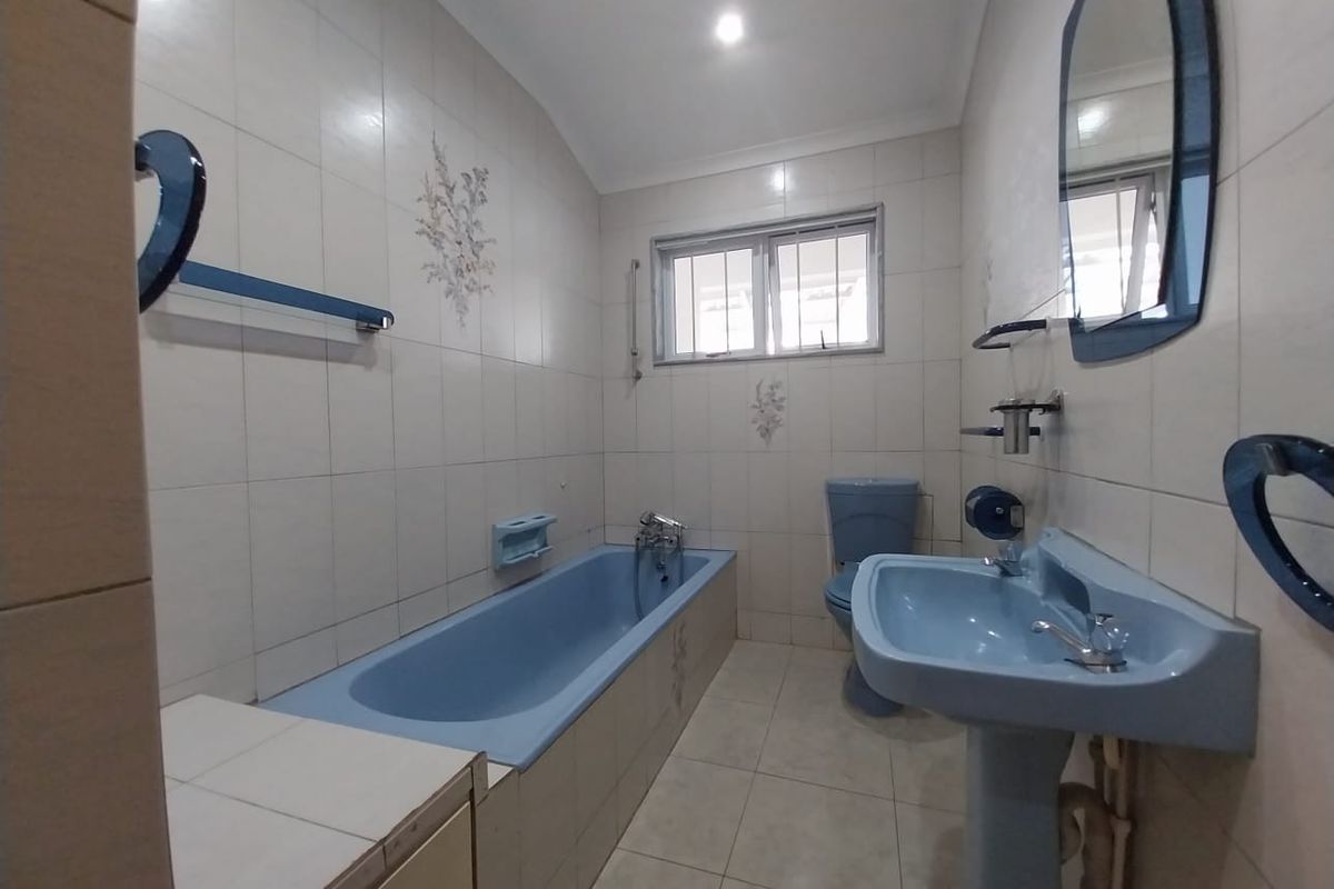 A dated bathroom featuring a blue bathtub, pedestal sink, and toilet, complemented by neutral wall tiles and a functional window for ventilation.