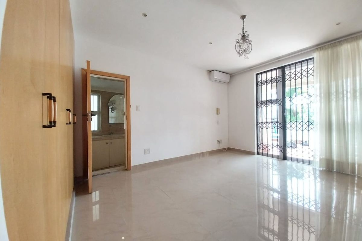 A bright bedroom featuring built-in wooden cupboards, tiled flooring, an en-suite bathroom, and a sliding door with security gate leading to the exterior.