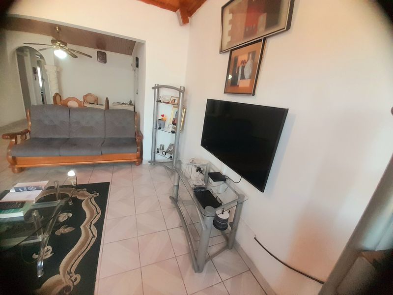 A living room featuring tiled flooring and exposed timber ceiling beams, with a wall-mounted television and an open-plan layout leading to an adjacent space.