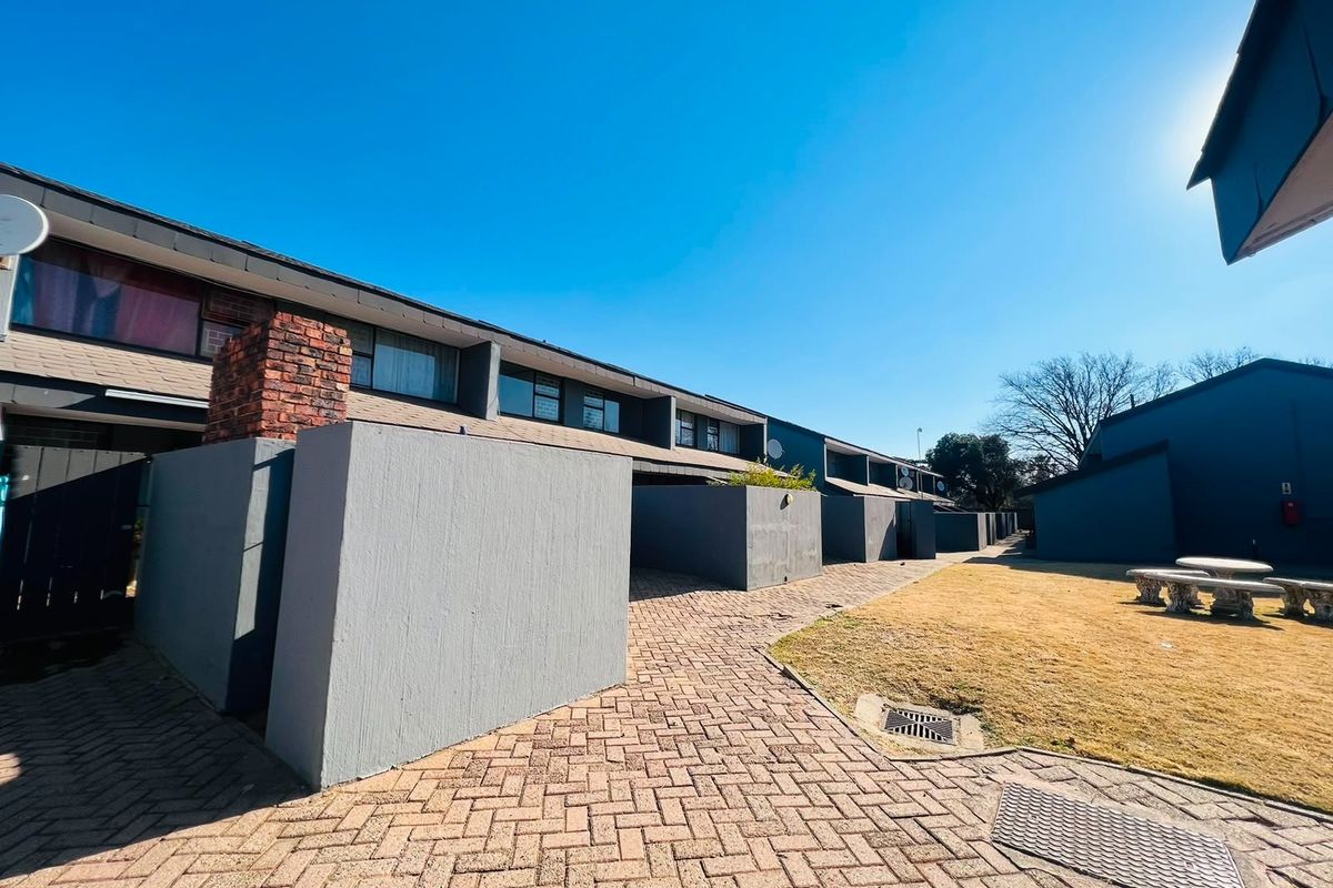 Exterior view of a townhouse complex featuring paved walkways, private courtyard walls, and a shared lawn area under a clear blue sky.