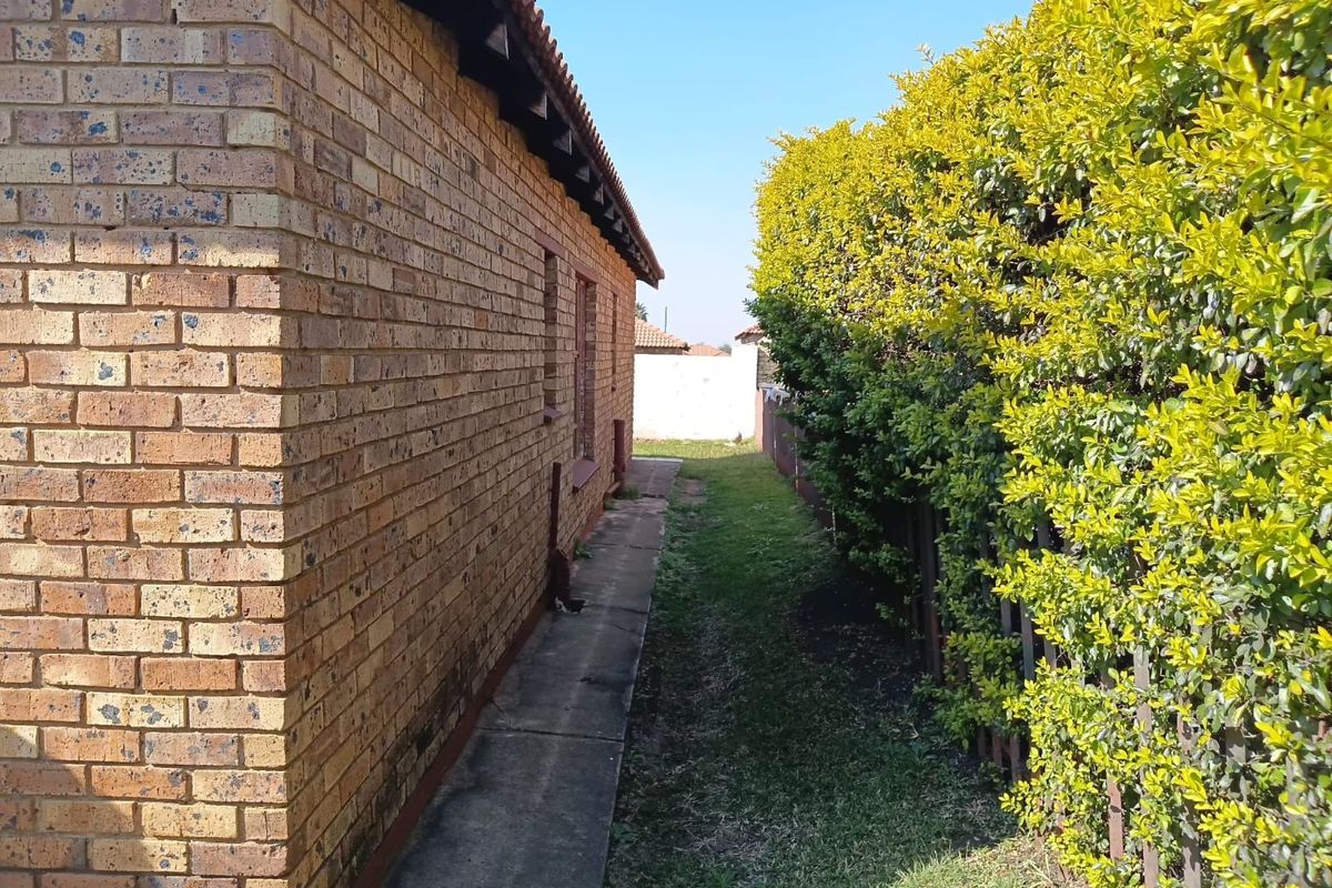 A side exterior view of the house featuring a brick facade, a concrete pathway, and a tall, established hedge along the property boundary.