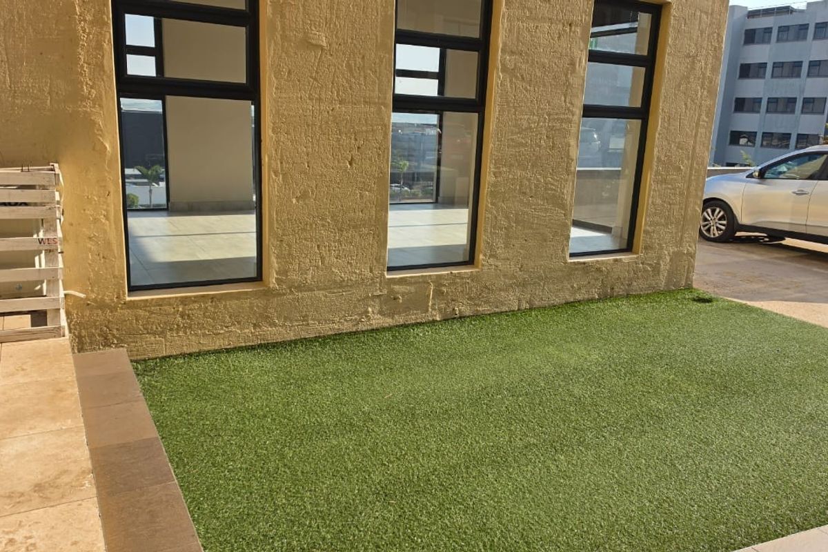 Exterior view of a commercial building featuring a small artificial grass lawn, paved patio area, and large glass windows with textured wall finish.