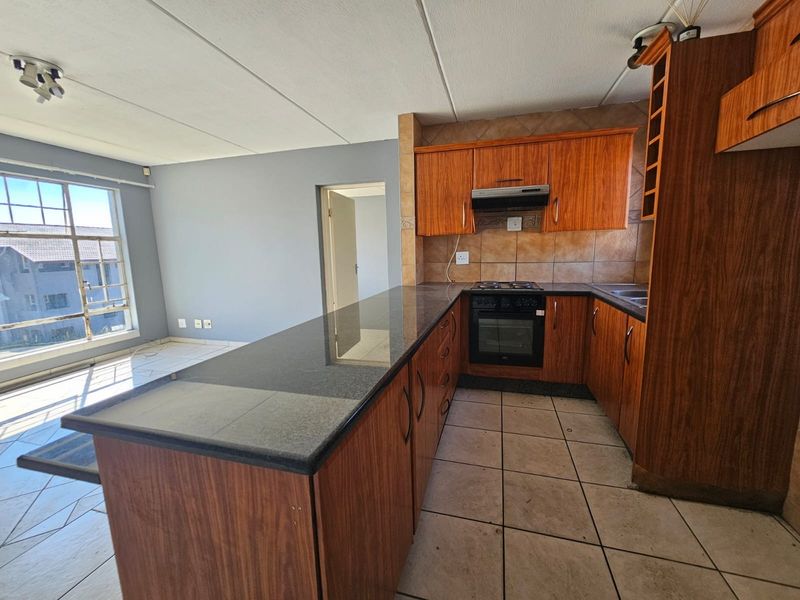 Open-plan kitchen featuring granite countertops, wooden cabinetry, and built-in appliances, flowing into a bright living area with large windows.
