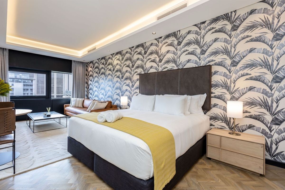 A modern, well-appointed bedroom featuring herringbone timber flooring, recessed ceiling lighting, and a striking feature wall with palm-leaf patterned wallpaper.