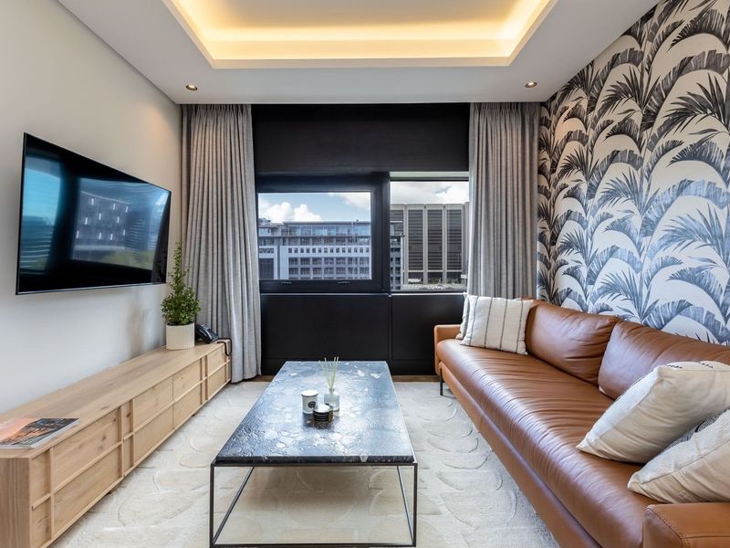 Modern living room featuring a recessed tray ceiling with integrated lighting, patterned feature wallpaper, and large windows offering an urban city view.
