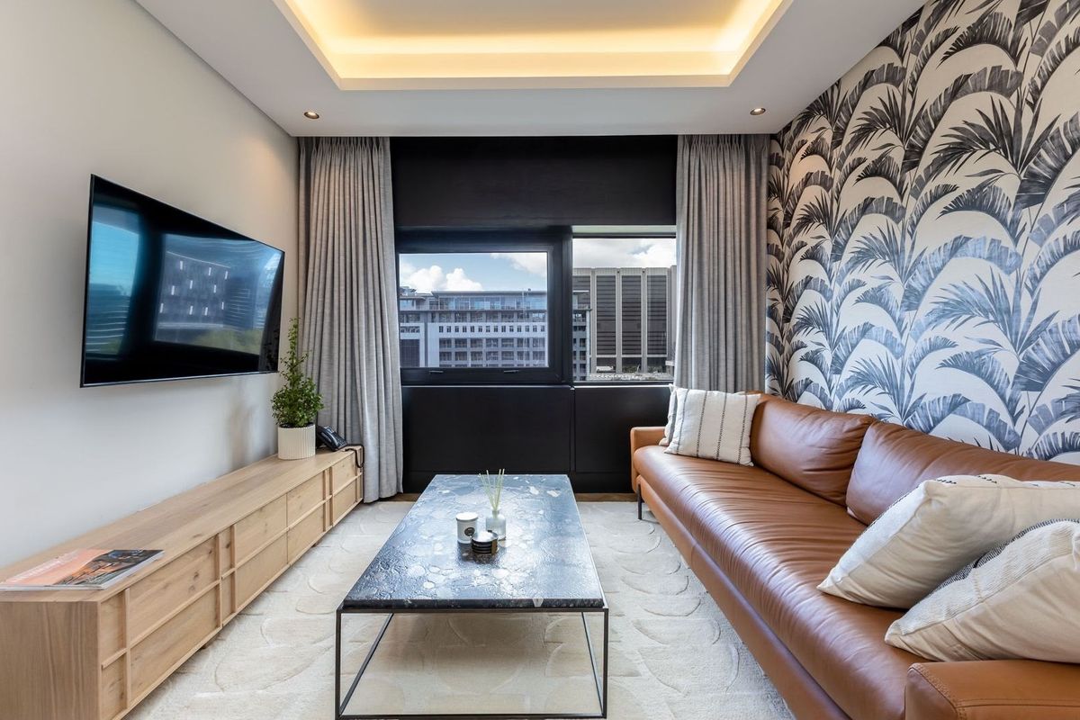 Modern living room featuring a recessed tray ceiling with integrated lighting, patterned feature wallpaper, and large windows offering an urban city view.