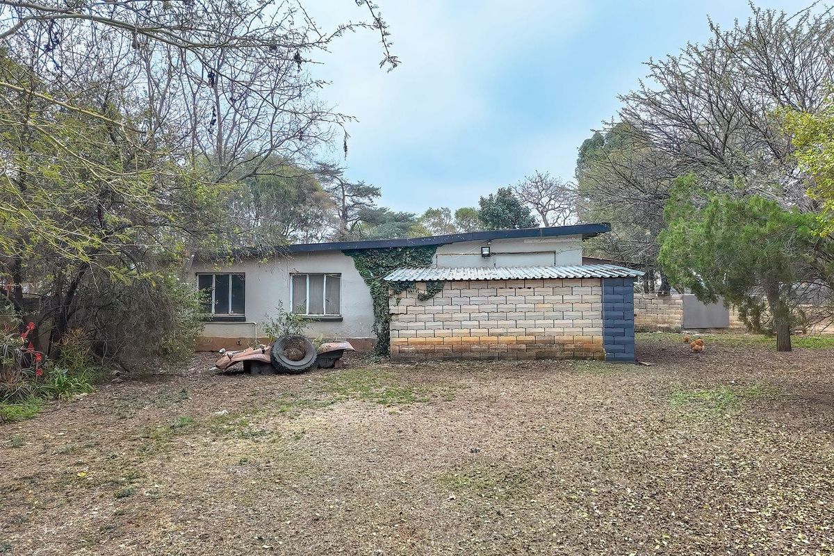 Rear view of a residential property featuring a single-story structure with a mix of masonry and plastered walls, set within a large, wooded garden area.