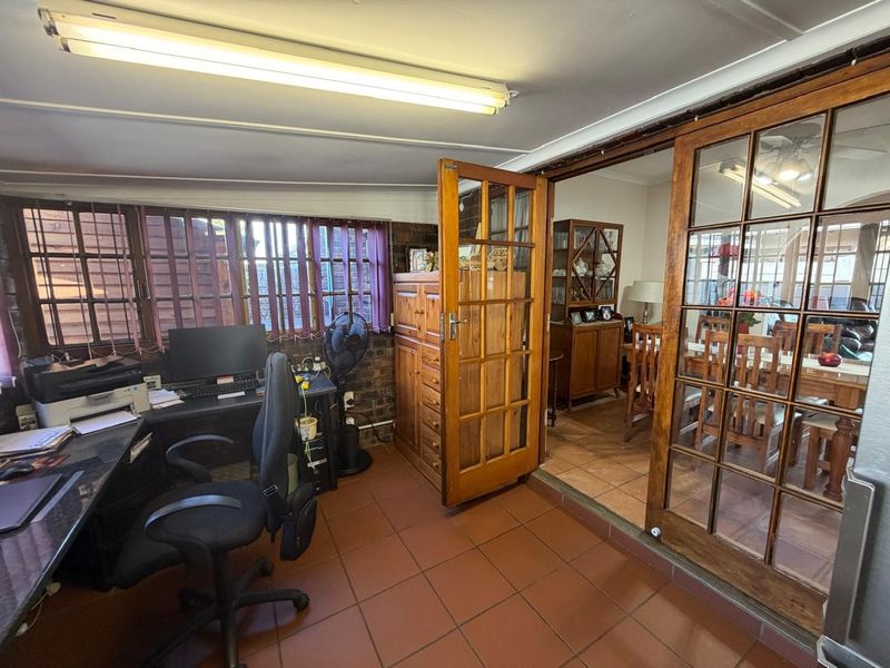 A functional home office space featuring terracotta tiled flooring, exposed brick accents, and wooden french doors connecting to the adjacent dining area.