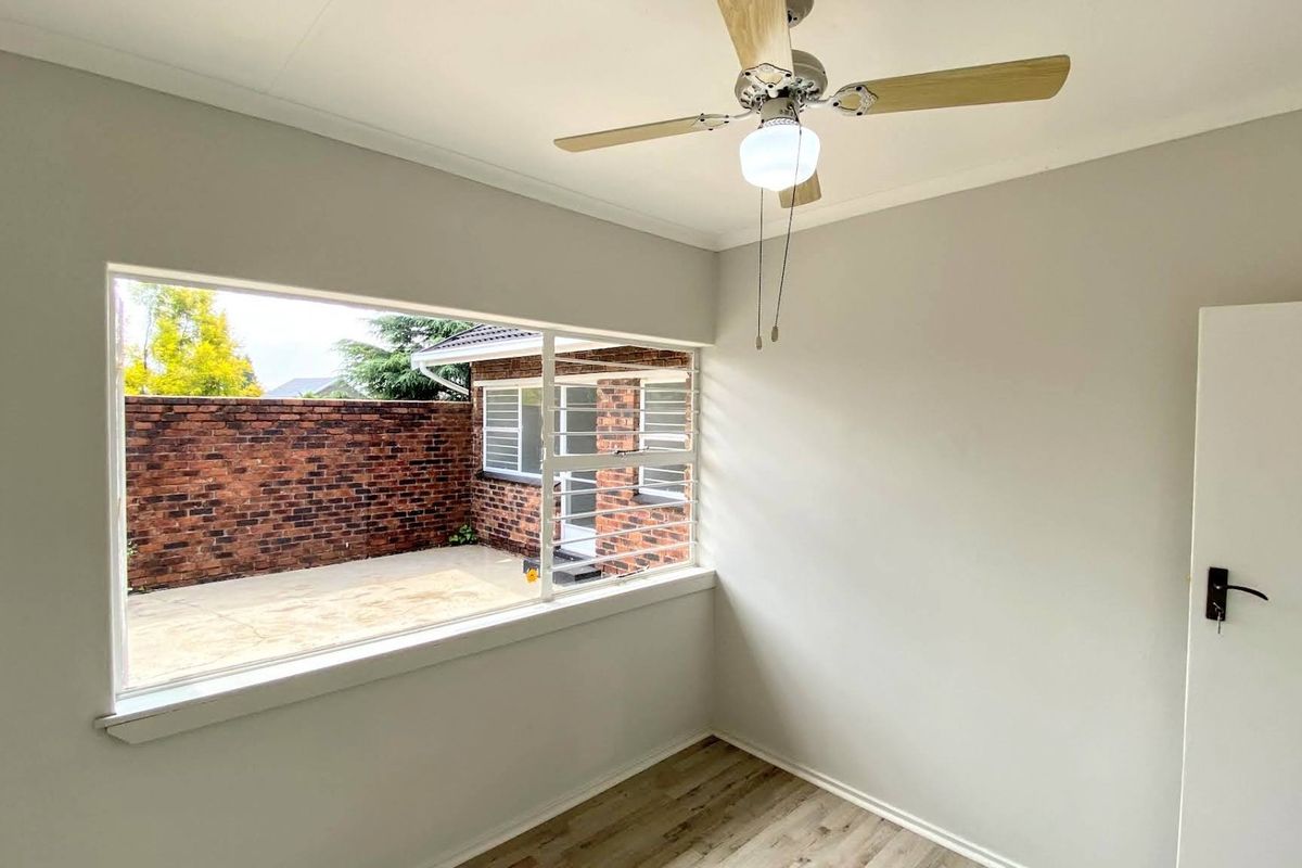 A bright, empty bedroom featuring light-toned laminate flooring, a ceiling fan, and a large window overlooking a private brick-walled courtyard.