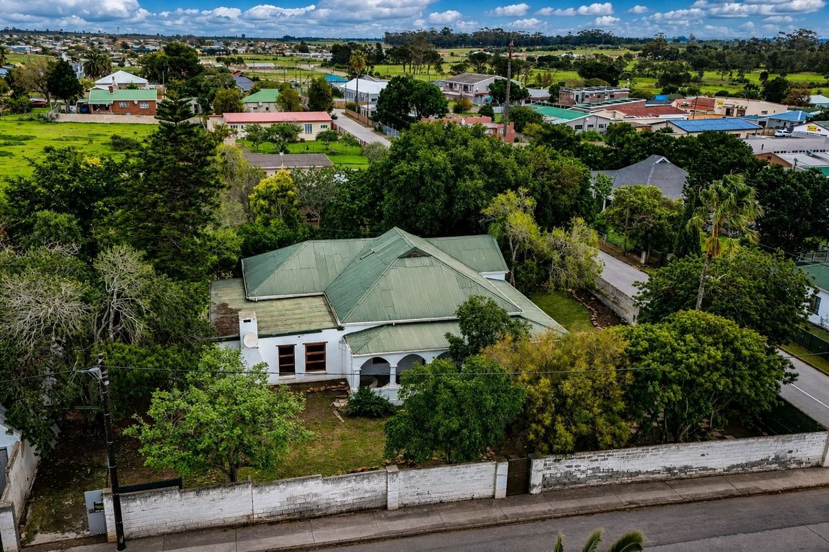 Aerial view of a residential property featuring a traditional house with a green metal roof, arched veranda, and a surrounding masonry boundary wall.