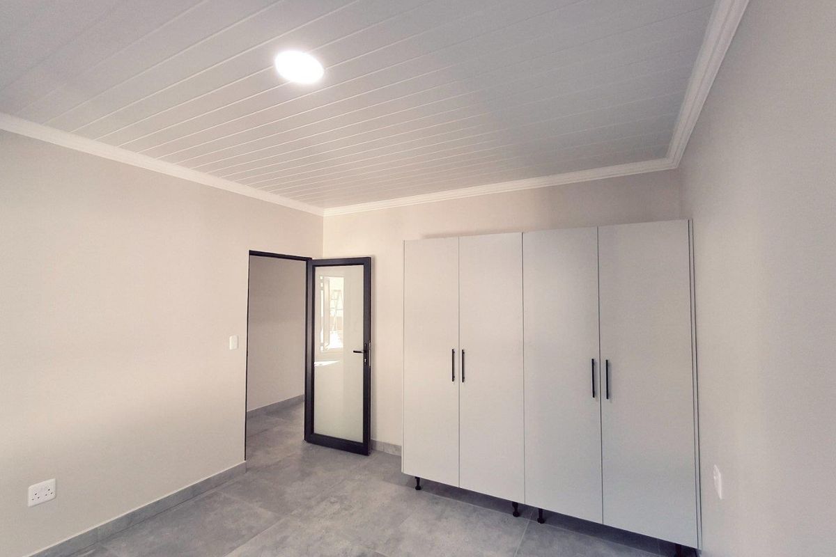 A clean, modern bedroom featuring neutral tiled flooring, a white tongue-and-groove ceiling, and built-in white cabinetry with sleek black handles.