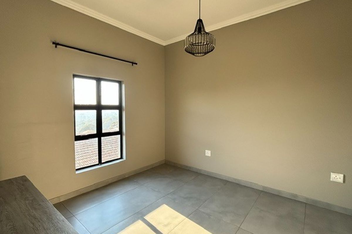 A bright, empty bedroom featuring neutral wall tones, large format floor tiles, crown molding, and a modern pendant light fixture.