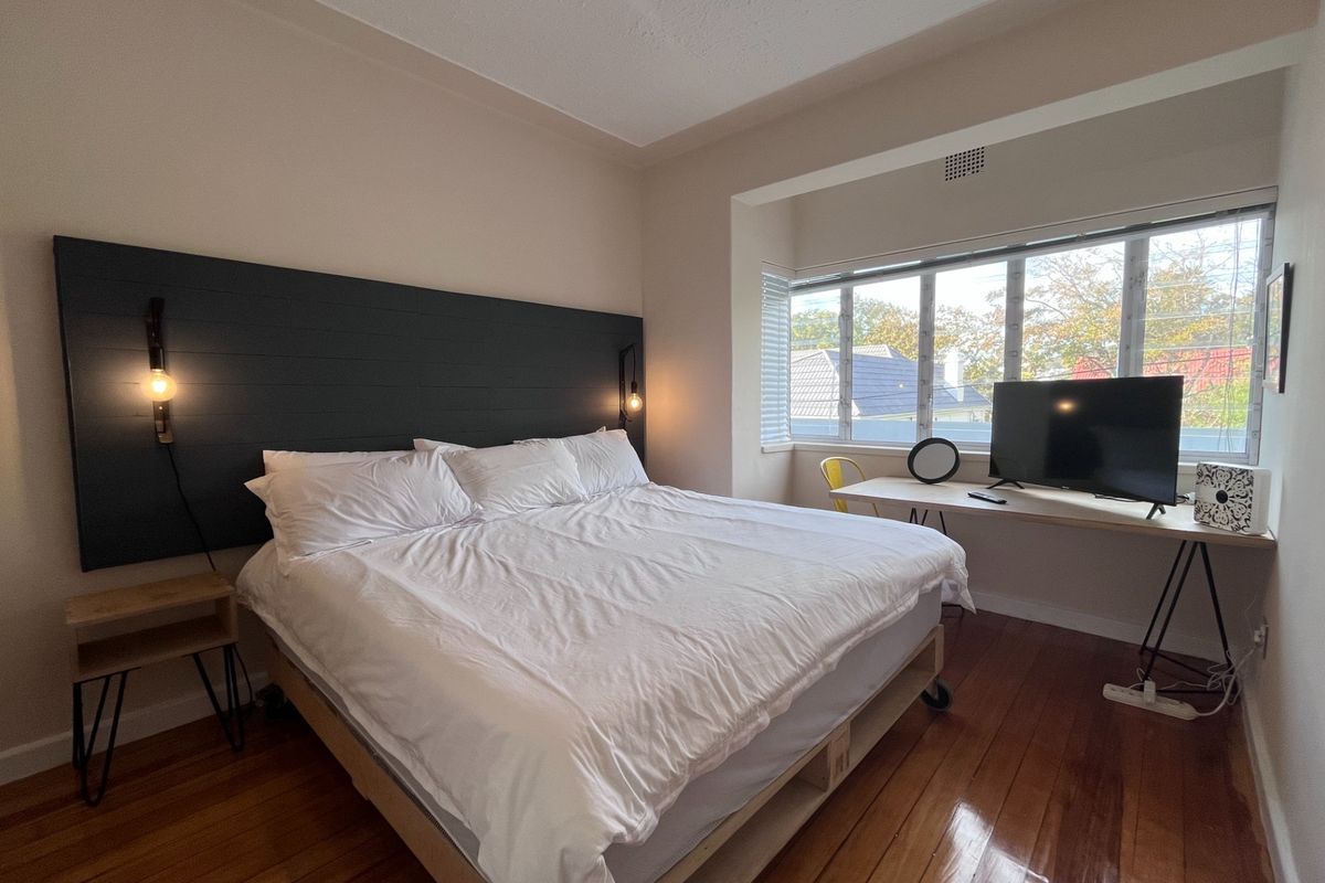 A well-lit bedroom featuring polished hardwood floors, a custom dark-toned headboard with integrated lighting, and a built-in desk area by the window.