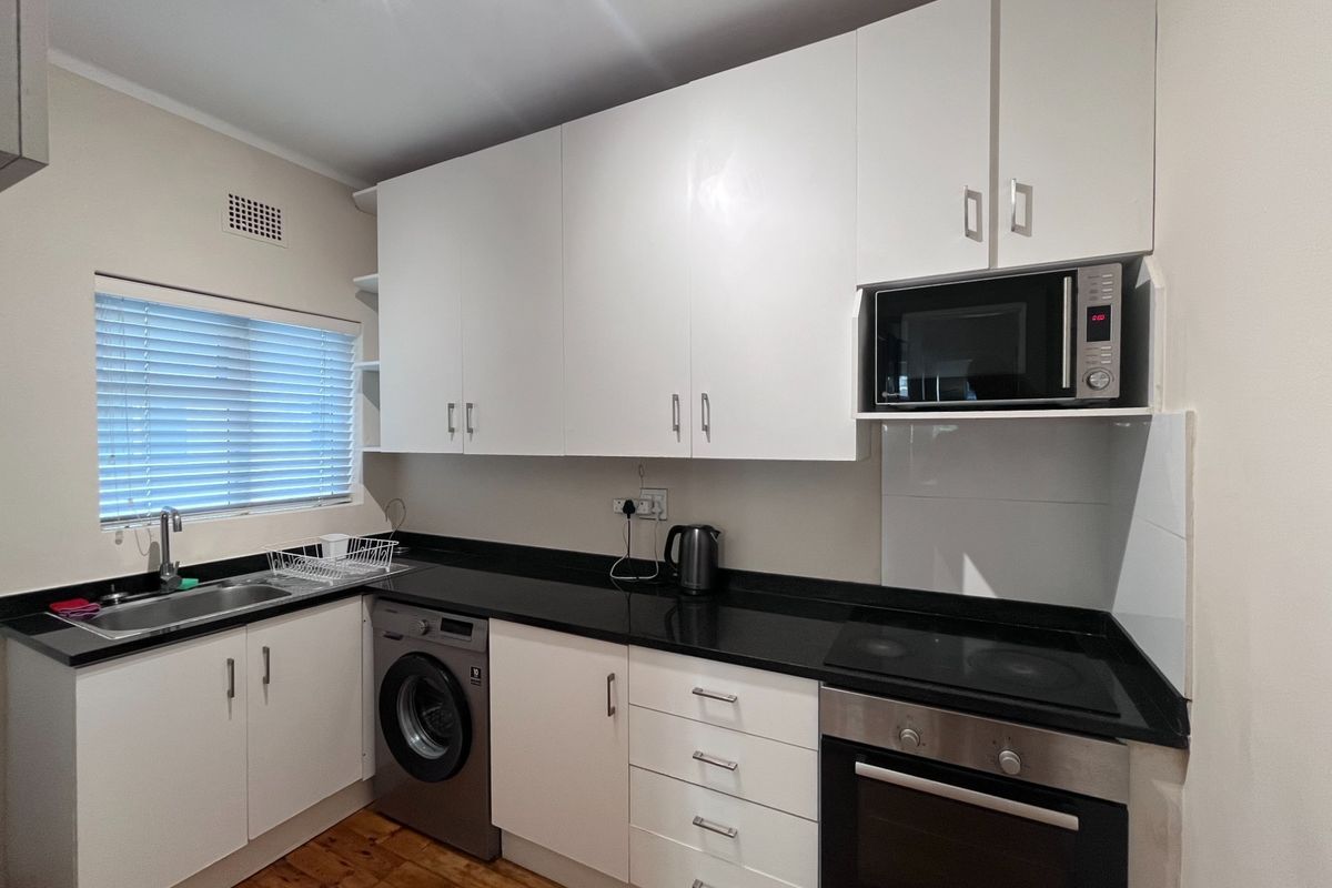 A functional kitchen featuring white cabinetry, black granite countertops, integrated oven, hob, and microwave, with a stainless steel sink and wooden flooring.