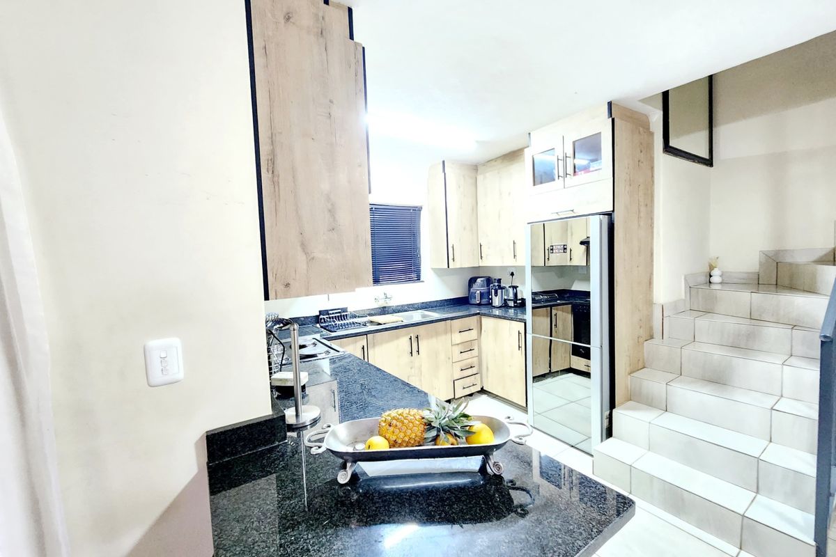 Modern kitchen featuring granite countertops, light wood-finish cabinetry, and integrated appliances, adjacent to a tiled staircase.