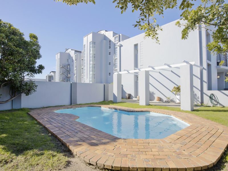 A private outdoor pool area featuring a brick-edged swimming pool, a pergola structure, and a surrounding lawn within a secure boundary wall.