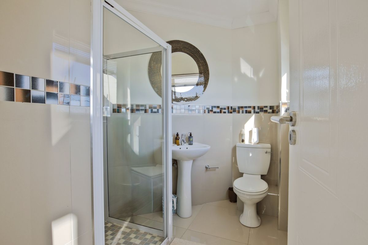 A functional bathroom featuring a pedestal basin, a glass-enclosed shower, and a toilet, finished with neutral wall tiles and a decorative mosaic border.