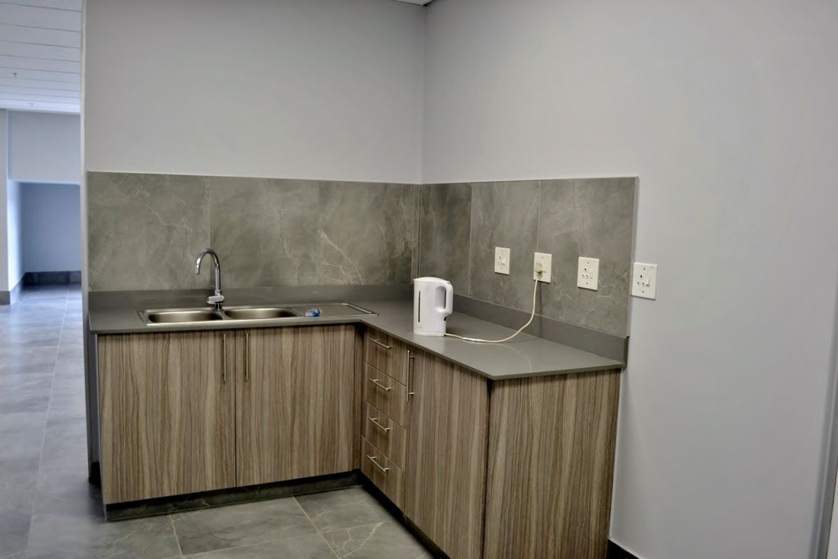 A clean, modern kitchenette area featuring laminate cabinetry, stone countertops, a stainless steel sink, and a tiled backsplash in an industrial office setting.