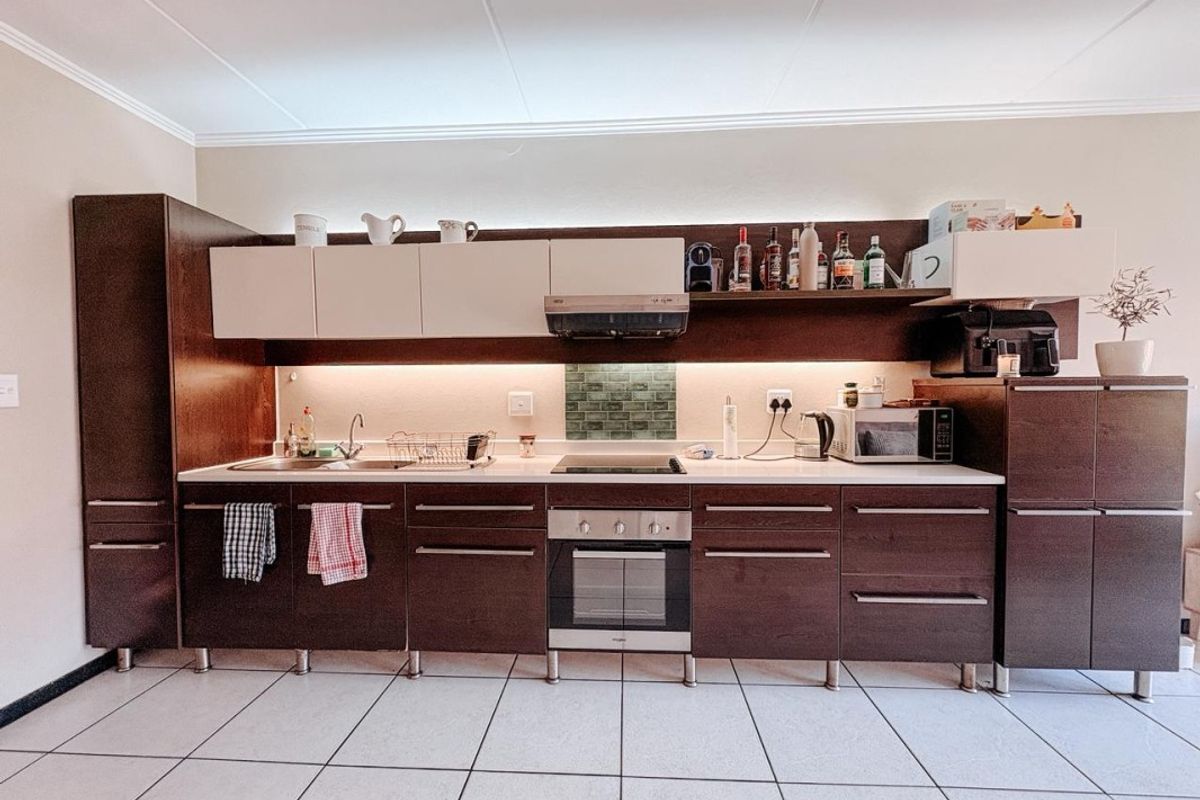 A modern kitchen featuring dark wood cabinetry, integrated oven and hob, under-cabinet lighting, and a tiled backsplash.