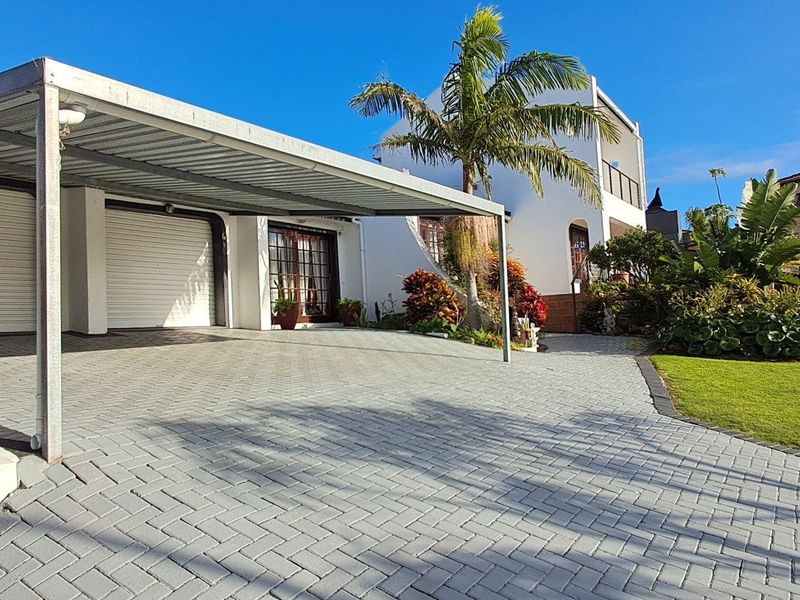 Front exterior view featuring a paved driveway, double garage with roller shutter doors, a covered carport, and a well-maintained landscaped garden.