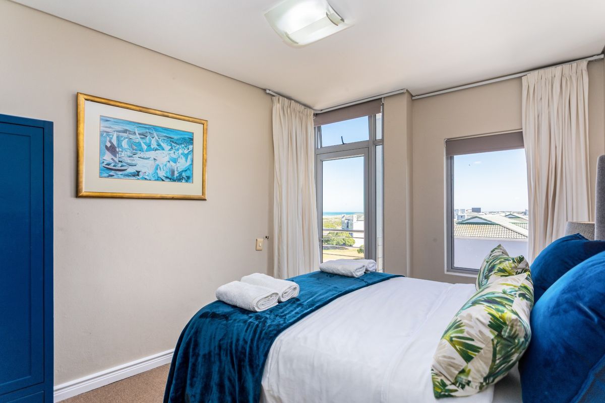 A bright bedroom featuring neutral walls, carpeted flooring, and large windows with access to a balcony, offering pleasant natural light and exterior views.
