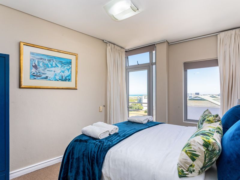 A bright bedroom featuring neutral walls, carpeted flooring, and large windows with access to a balcony, offering pleasant natural light and exterior views.