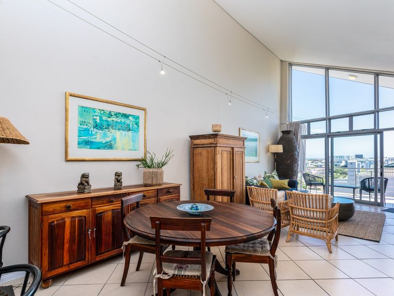 Spacious open-plan living and dining area featuring high vaulted ceilings, large floor-to-ceiling windows, and direct access to a private balcony with coastal views.