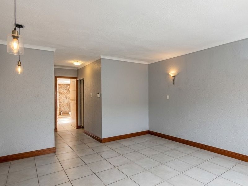 An empty, neutrally painted living area featuring tiled flooring, coving, and modern wall and pendant lighting, with a view into an adjacent bathroom.