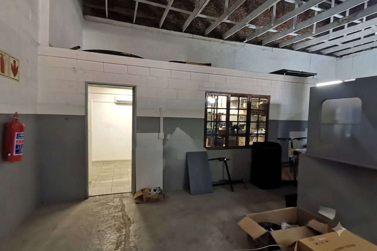 Industrial workspace featuring exposed ceiling rafters, concrete flooring, a partitioned office area with a window, and a wall-mounted fire extinguisher.