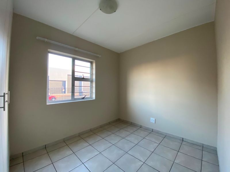 A clean, empty bedroom featuring neutral wall paint, tiled flooring, a built-in cupboard, and a window fitted with security bars.