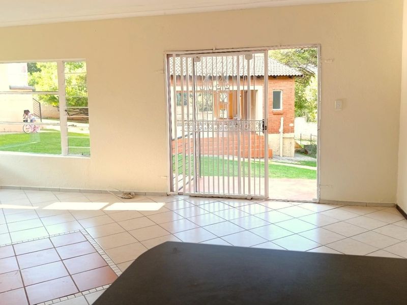 A bright, empty living area featuring tiled flooring, large windows, and a sliding security gate providing access to the outdoor garden space.