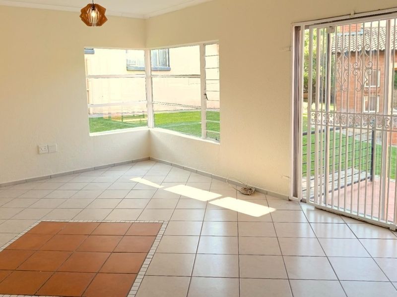 An empty, sunlit living area featuring neutral tiled flooring, corner windows, and a sliding door leading to an outdoor patio space.