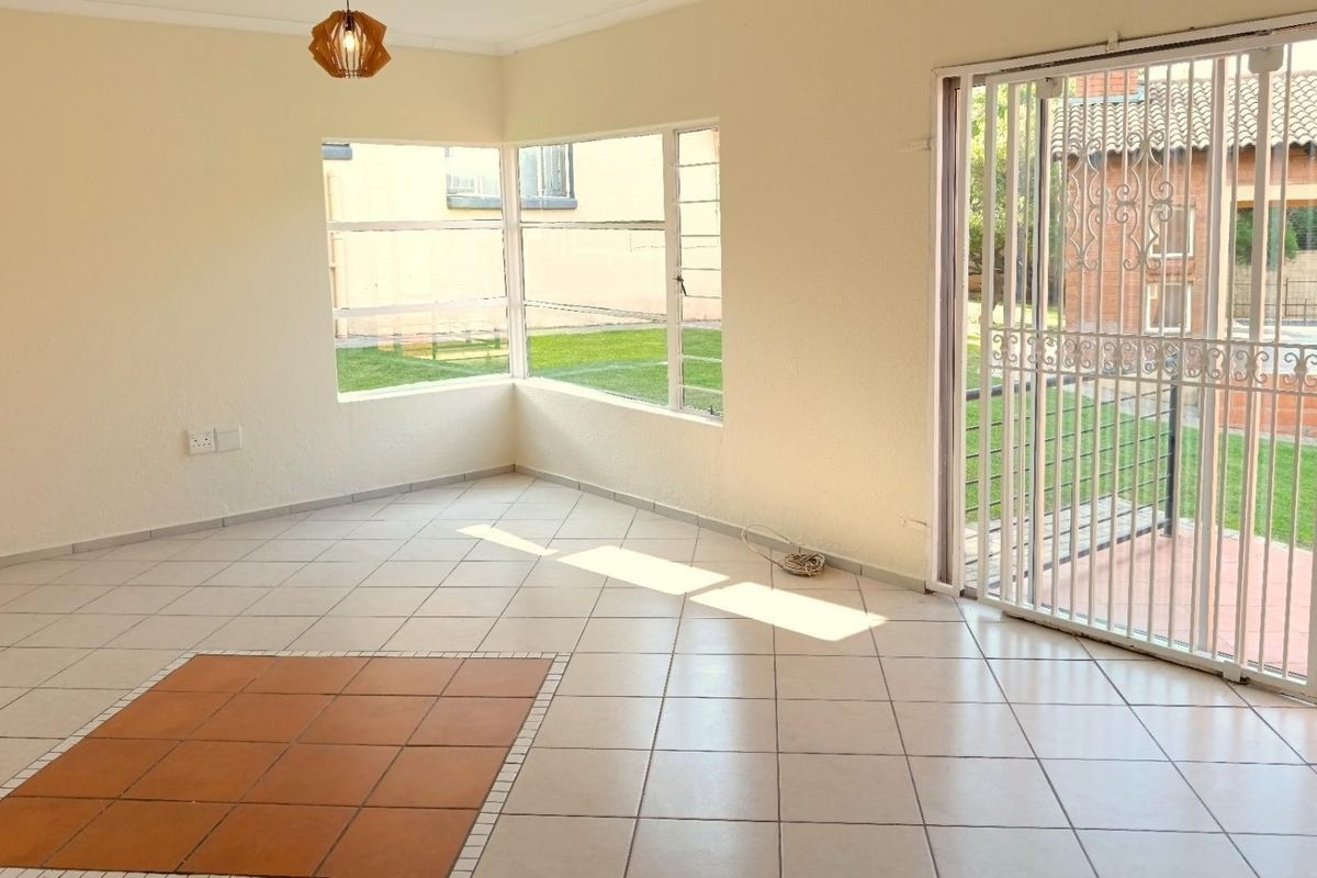 An empty, sunlit living area featuring neutral tiled flooring, corner windows, and a sliding door leading to an outdoor patio space.