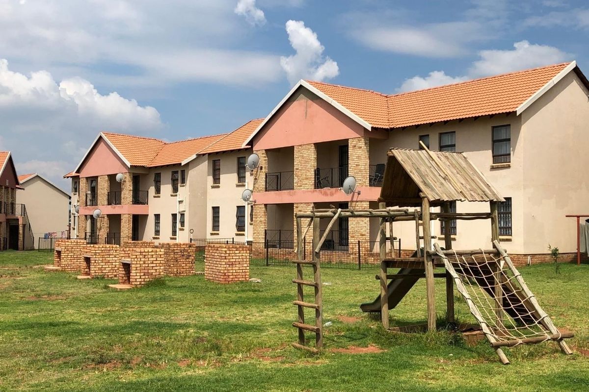 Exterior view of an apartment complex featuring communal brick braai areas and a playground set within a shared garden space.