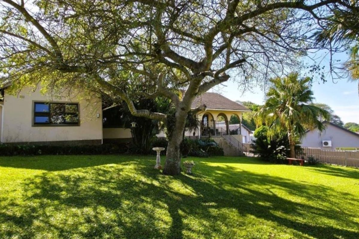 A spacious rear garden featuring a well-maintained lawn, mature trees, and a view of the house exterior with a covered patio area.