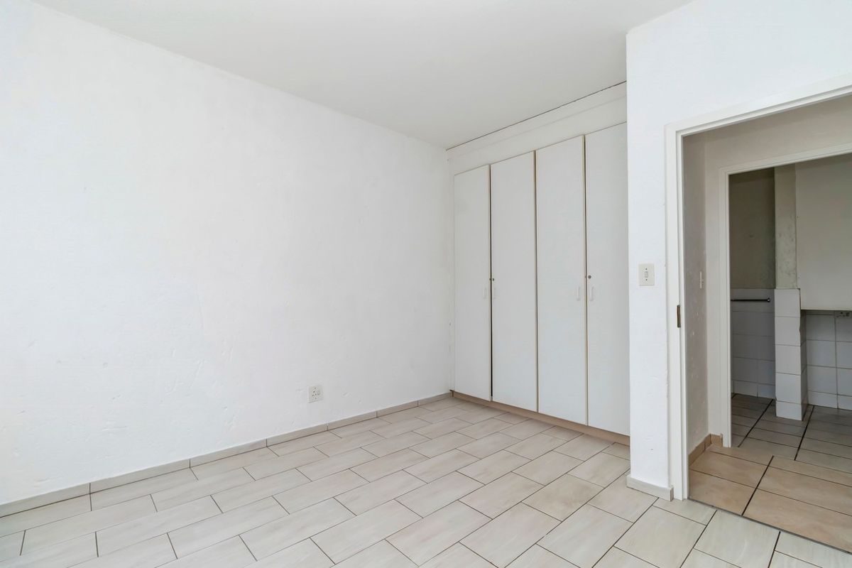 A bright, empty bedroom featuring neutral floor tiling and built-in white cabinetry, with an adjacent doorway leading to a tiled utility or bathroom area.