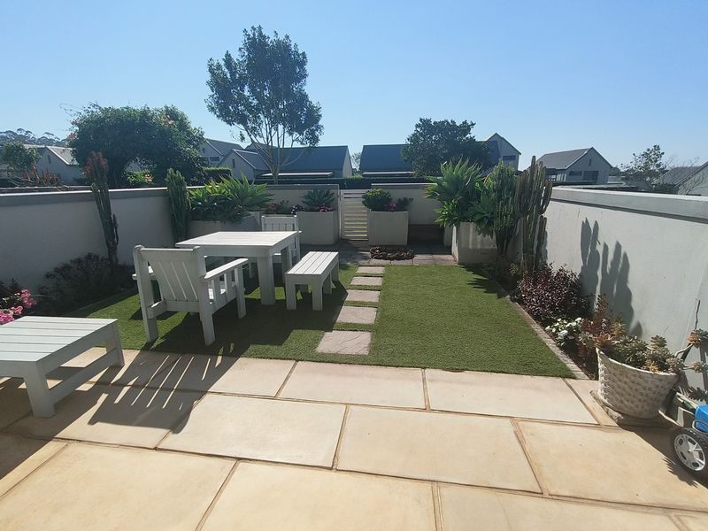 A well-maintained rear courtyard featuring a paved patio area, artificial lawn, stone pathway, and integrated boundary wall planters.
