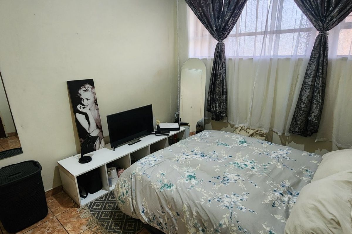 A compact bedroom featuring tiled flooring and a window with security bars, offering a functional space with potential for cosmetic updates.